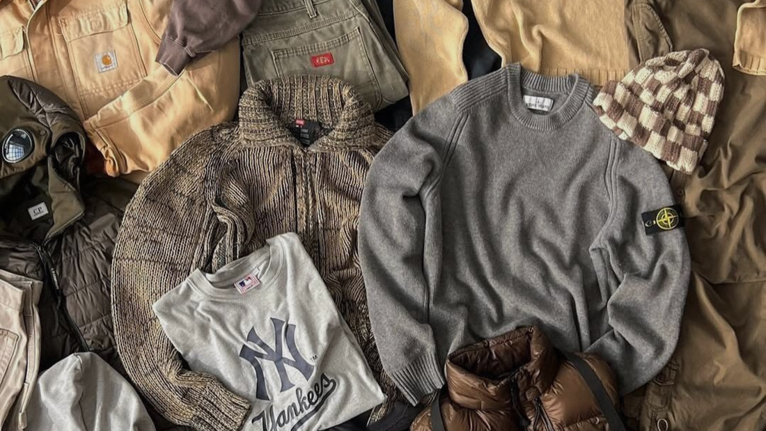 Composizione di abbigliamento streetwear invernale da uomo con maglione grigio Stone Island, giacca senape Carhartt, t-shirt grigia New York Yankees, giubbotto verde C.P. Company con lenti, pantaloni cargo e berretto a scacchi.