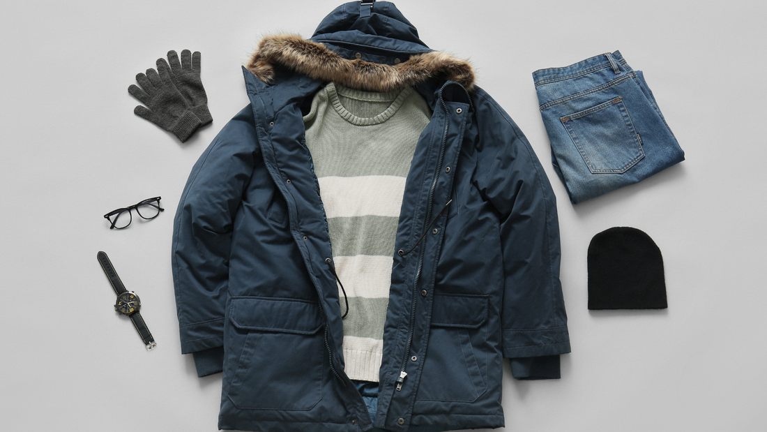 Completo invernale piatto comprensivo di un parka blu scuro con bordo in pelliccia sintetica, un maglione a righe verde e panna, guanti grigi, occhiali, un orologio, jeans e un berretto nero su uno sfondo grigio.