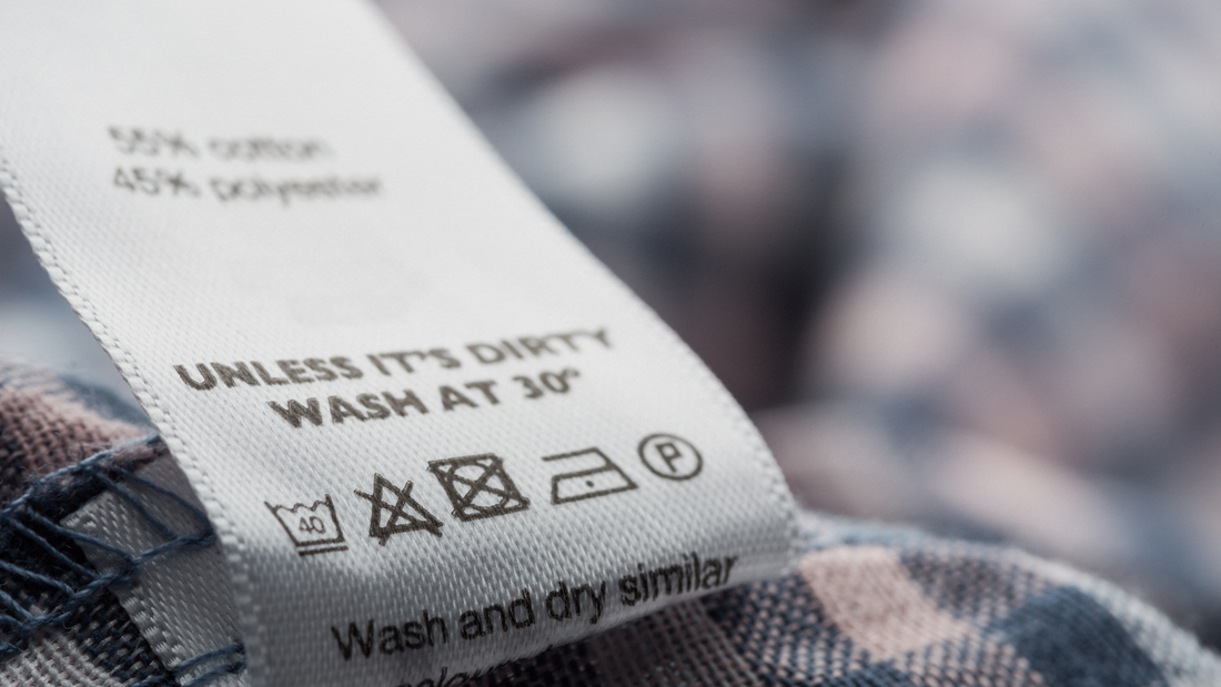 Dettaglio di un'etichetta di lavaggio cucita su un tessuto a quadri, con indicata la composizione (55% cotone, 45% poliestere), il testo "UNLESS IT'S DIRTY WASH AT 30°" e i simboli per la cura del capo.