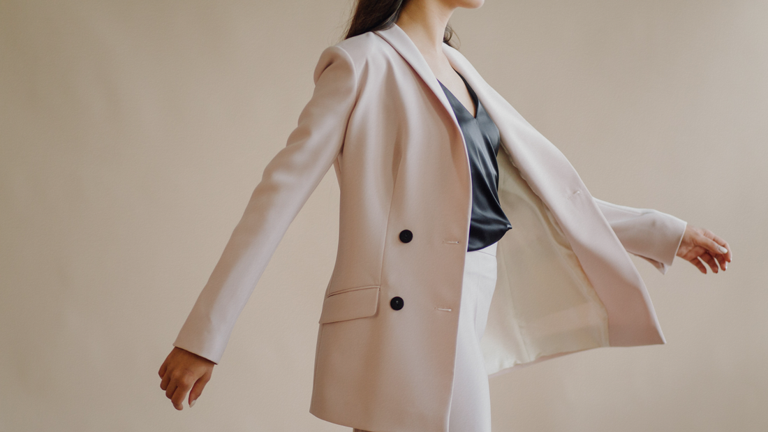 Giacca blazer doppiopetto rosa cipria da donna aperta e fluttuante con bottoni neri a contrasto, top scuro in raso e pantaloni coordinati chiari.