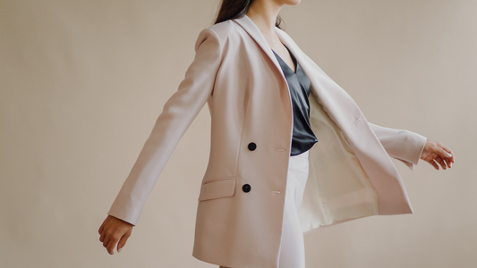 Giacca blazer doppiopetto rosa cipria da donna aperta e fluttuante con bottoni neri a contrasto, top scuro in raso e pantaloni coordinati chiari.