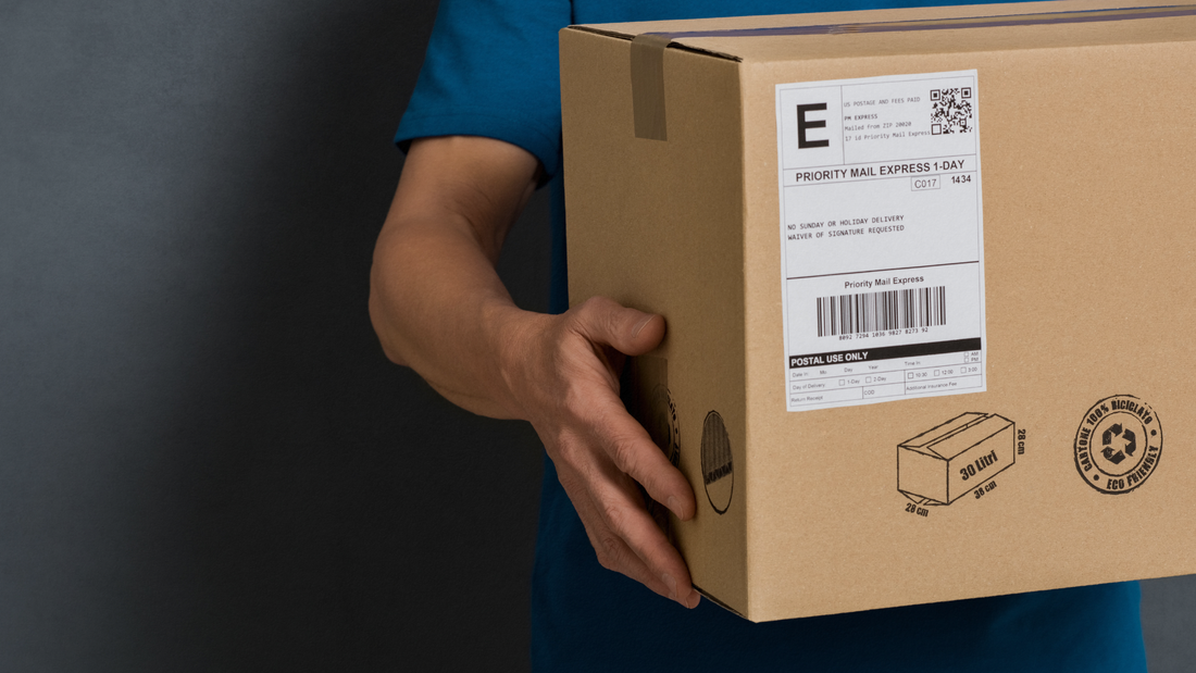 Dettaglio di una persona con maglietta blu che tiene in mano un pacco da spedizione in cartone con etichetta Priority Mail Express e simbolo del cartone riciclato.