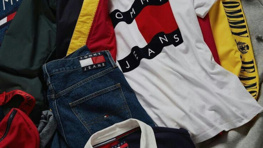 Composizione di abbigliamento Tommy Jeans e Tommy Hilfiger con t-shirt bianca con maxi logo, pantaloni in denim blu con patch, polo blu scuro con colletto bianco e altri capi sportivi nei colori rosso, giallo e blu.