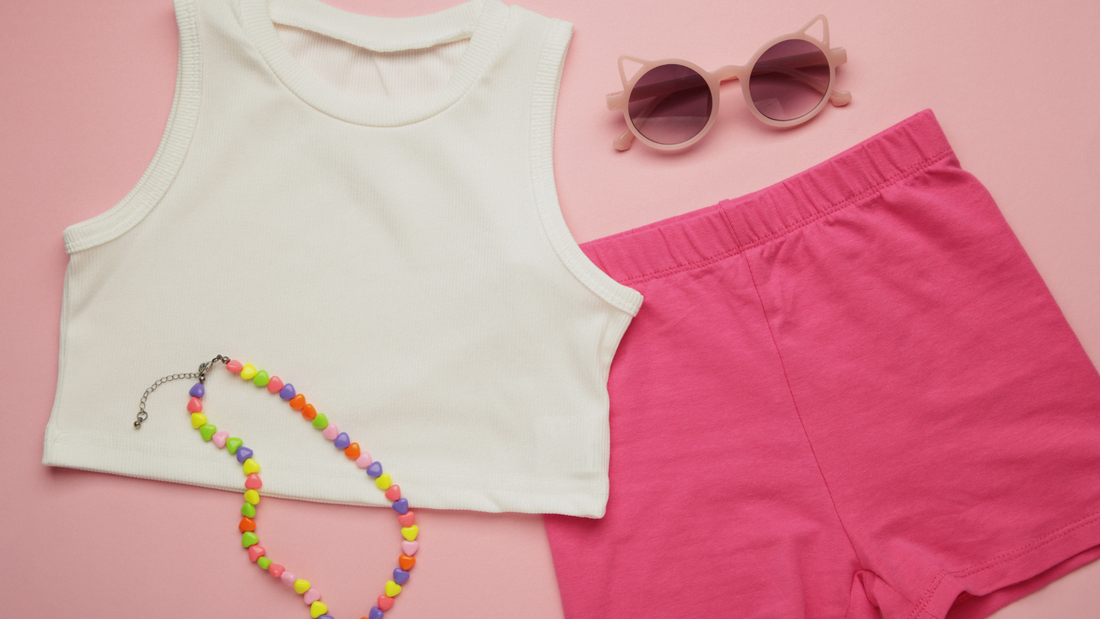Flat lay di un outfit estivo da bambina con crop top bianco a coste, pantaloncini da ciclista rosa acceso, collana di perline colorate a cuore e occhiali da sole rosa, su sfondo rosa pastello.